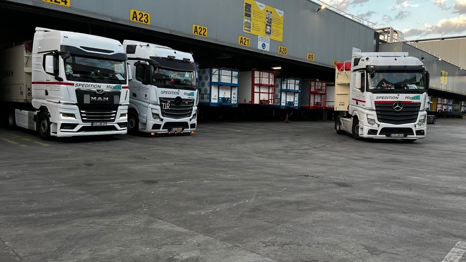 Drei weiße Sattelschlepper parken an Laderampen, ein vierter Lastwagen fährt ein, alles in einem Industriegebiet.