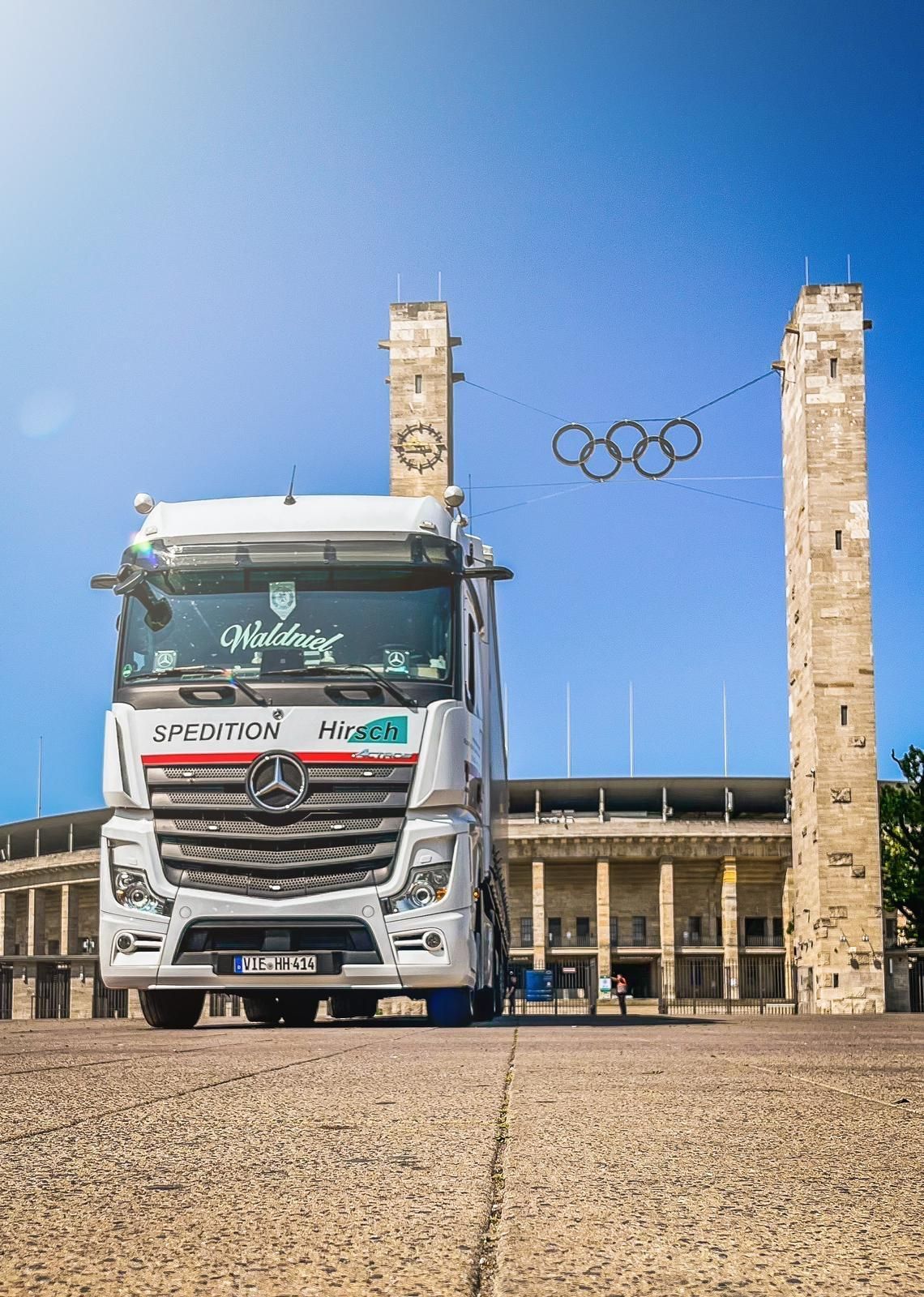 Weißer LKW vor dem Berliner Olympiastadion. Über dem Kopf hängen Olympische Ringe. Blauer Himmel.