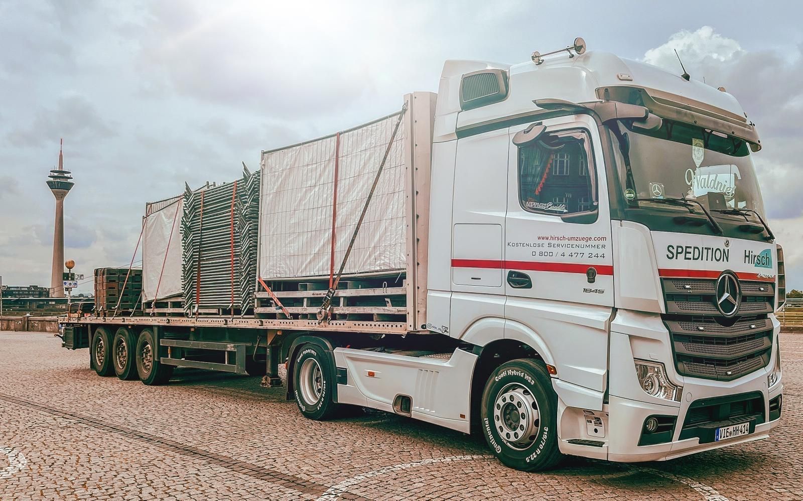 Ein weißer Mercedes-LKW mit einem Pritschenanhänger, der Ladung transportiert; im Hintergrund ist der Düsseldorfer Fernsehturm zu sehen.