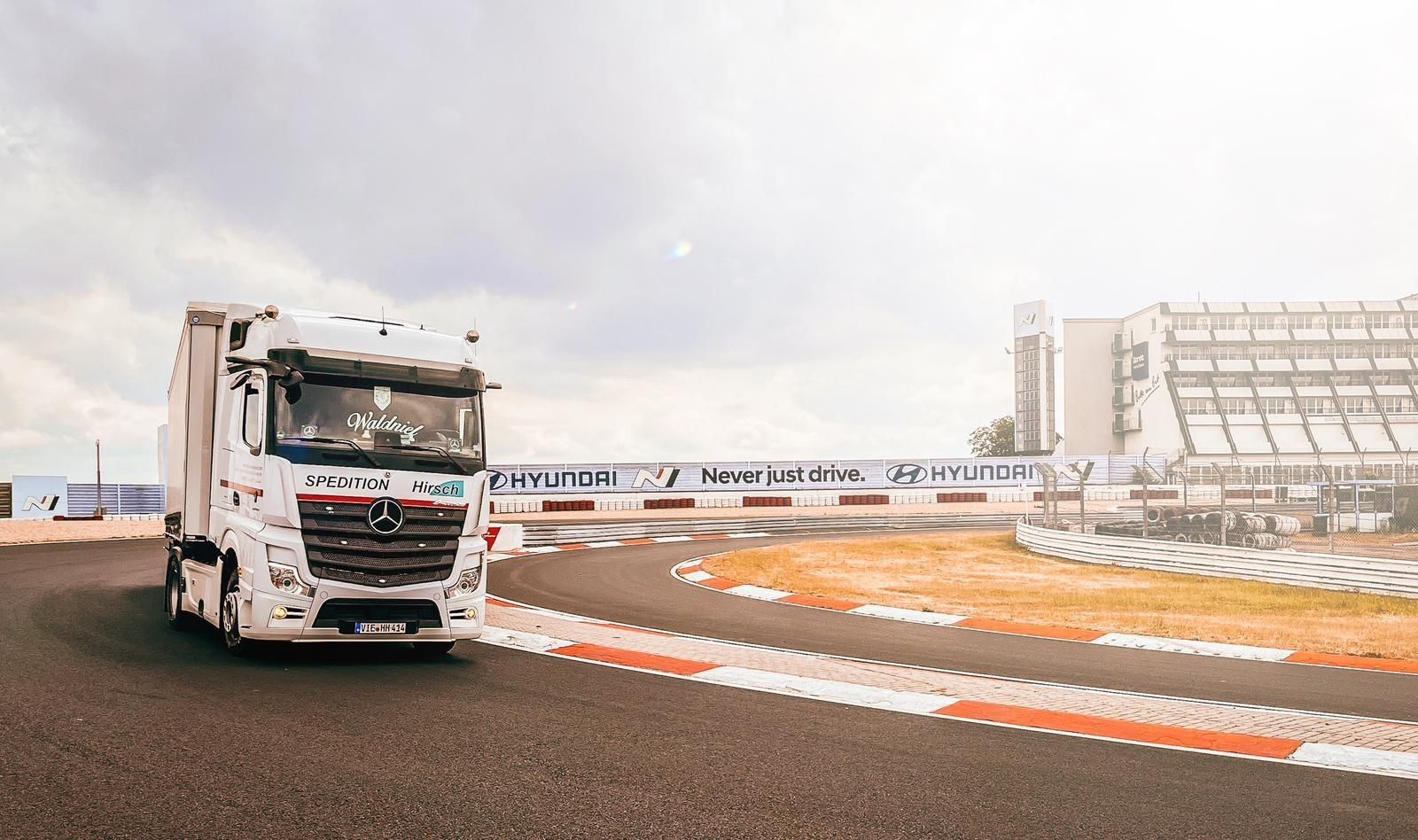 Ein weißer Mercedes-Truck auf einer Rennstrecke; im Hintergrund sind ein Tor und eine Tribüne zu sehen.
