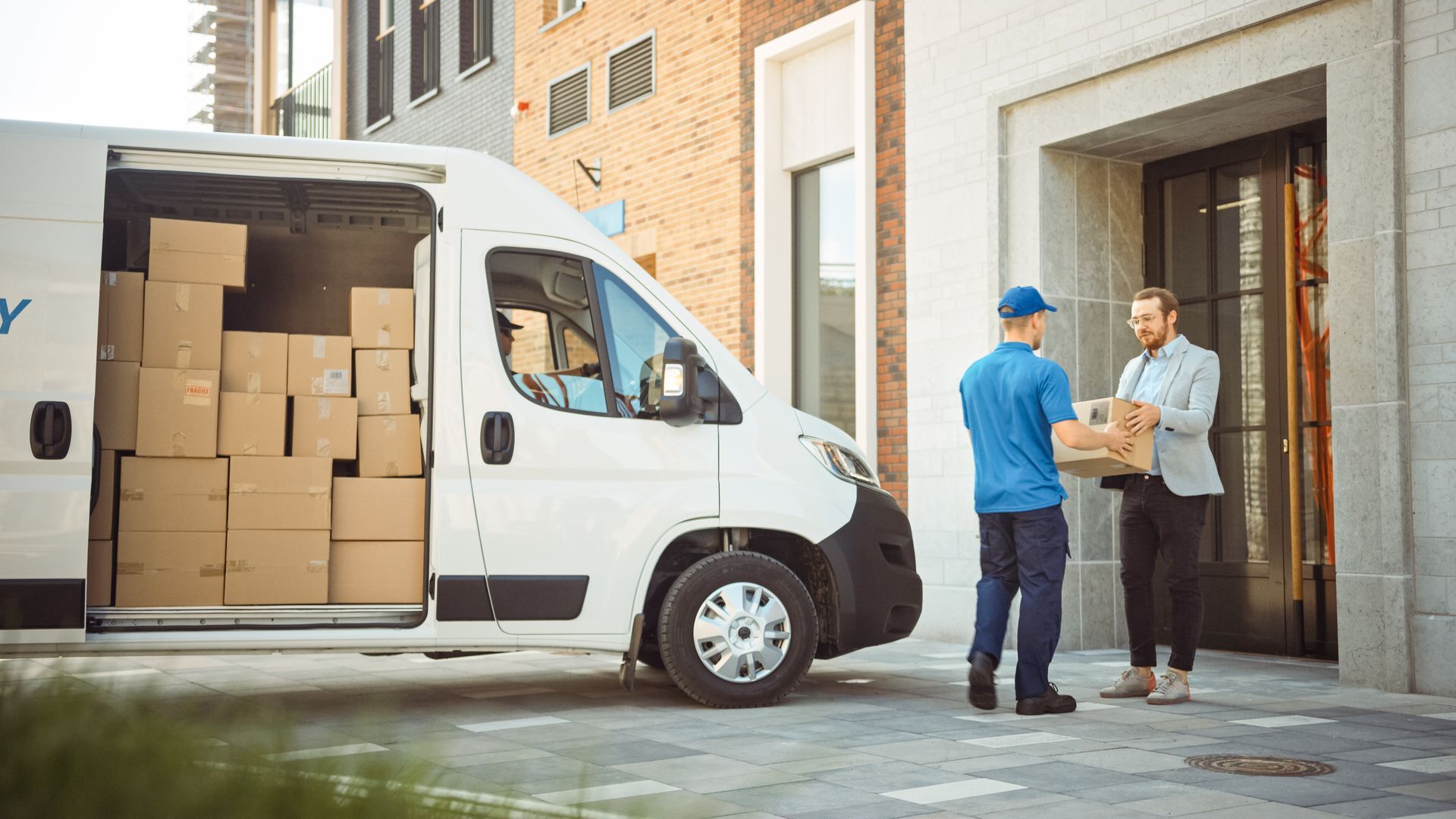 Un livreur remet un colis à un client devant un bâtiment. Une camionnette blanche, des cartons à l'intérieur.