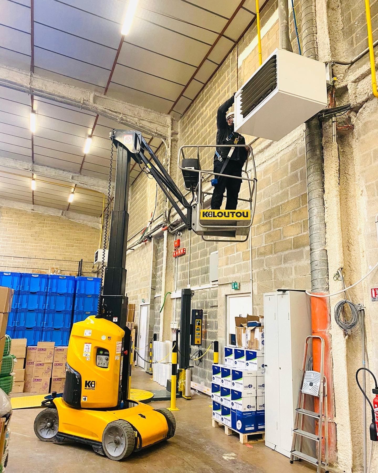 Un technicien, installé dans une nacelle élévatrice, effectue la maintenance d'un système de climatisation.