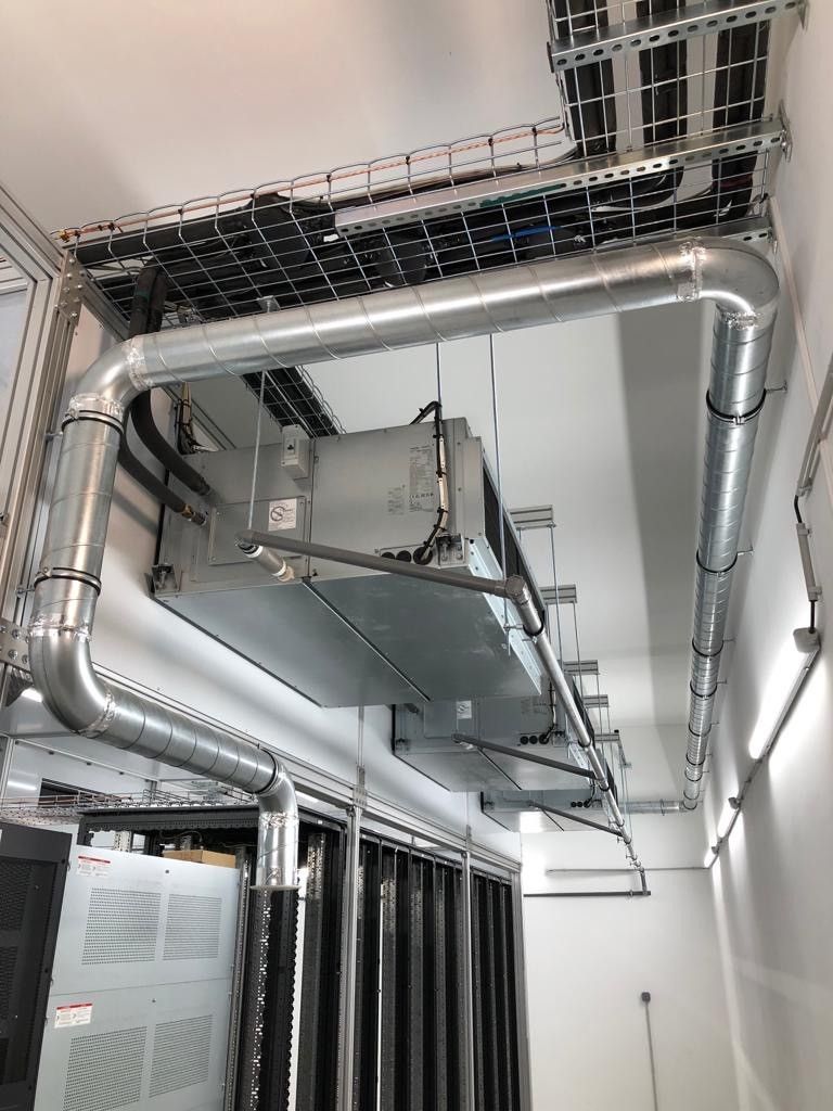 Des conduits de ventilation sont suspendus à un plafond au-dessus des baies de serveurs dans une salle technique.