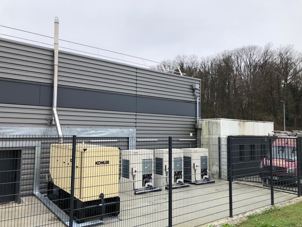 Un générateur industriel beige et plusieurs unités de refroidissement sont installés à l'extérieur d'un bâtiment métallique.