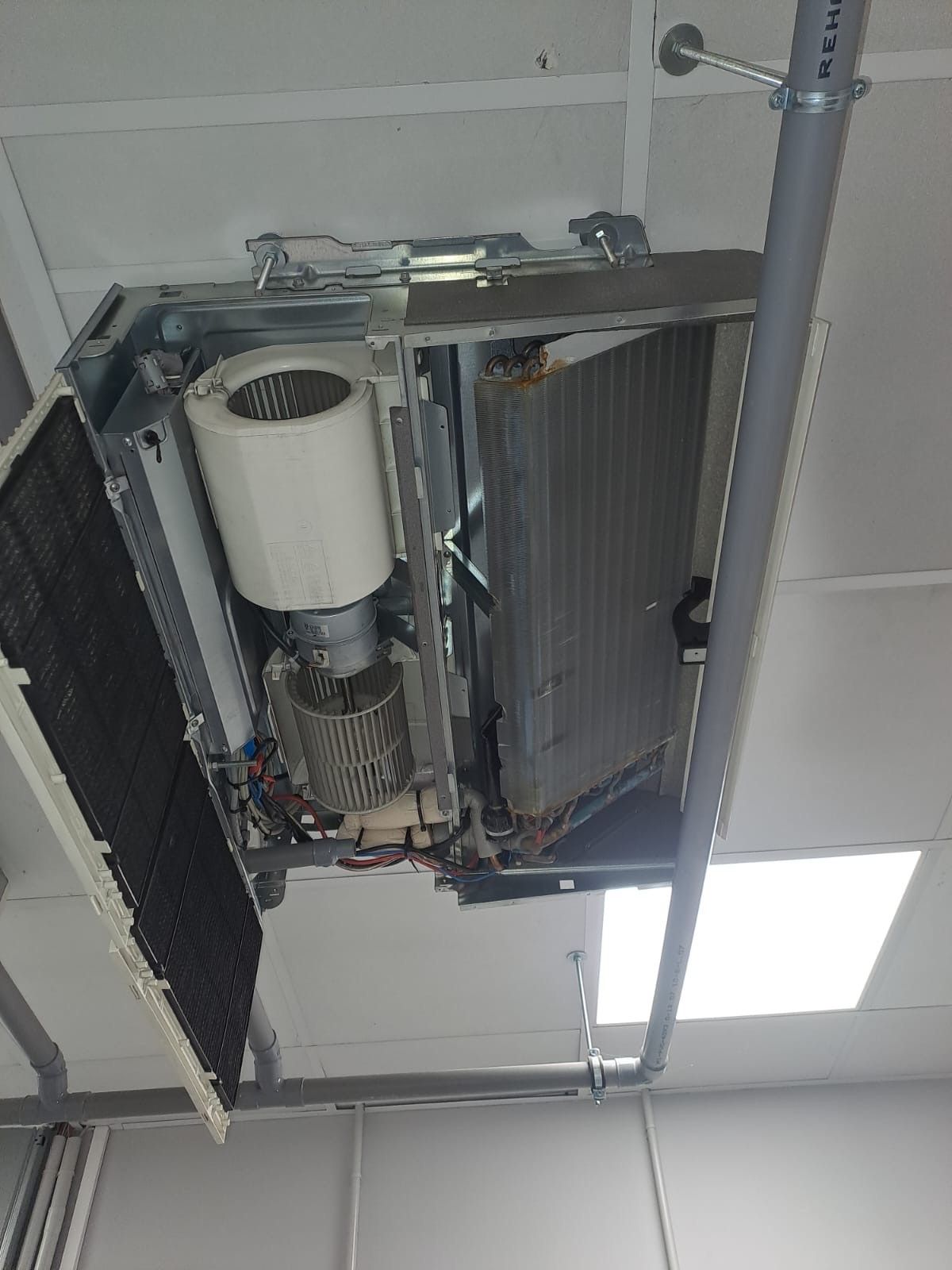 Un climatiseur apparent, fixé au plafond, avec un ventilateur et des serpentins métalliques visibles.