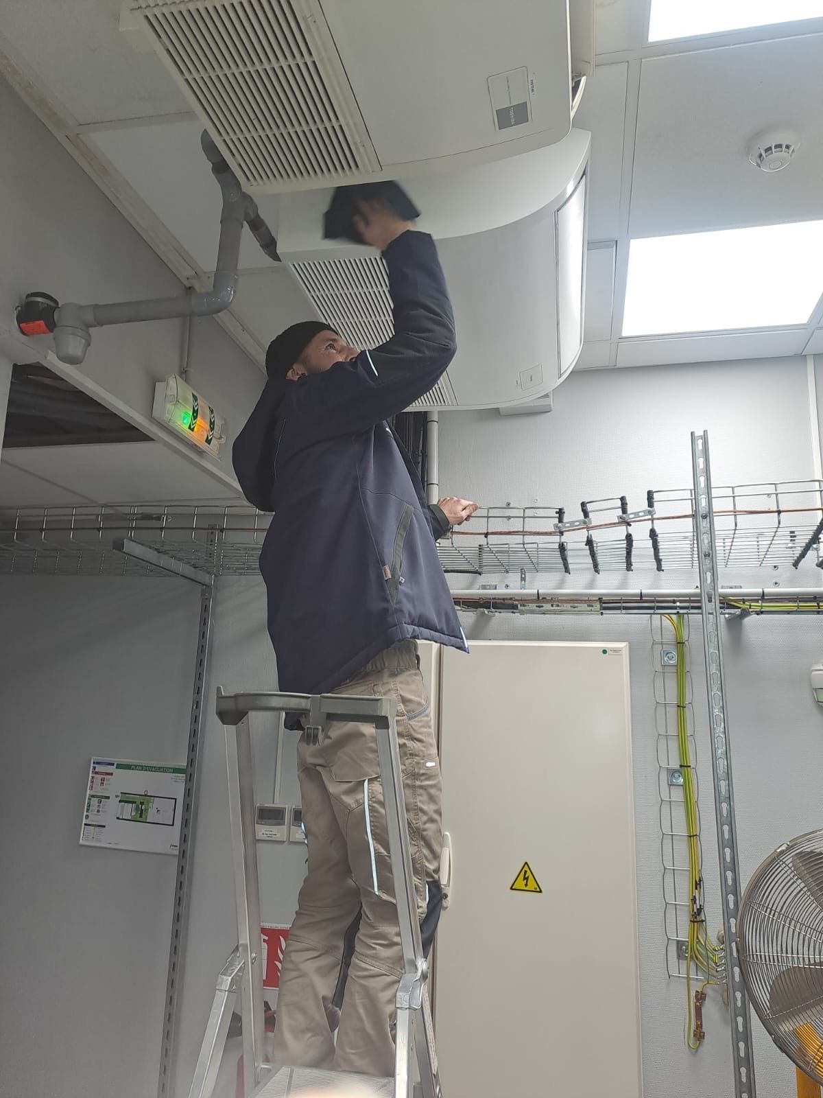 Une personne debout sur une échelle effectue la maintenance d'un climatiseur fixé au plafond dans un local technique.