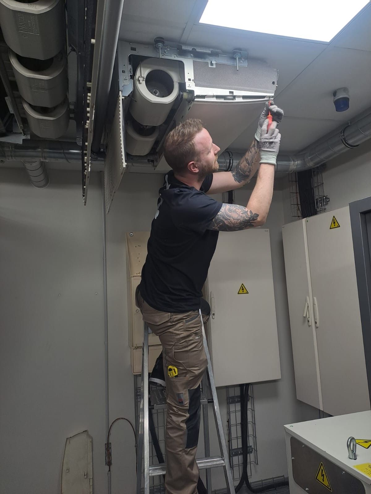 Un technicien, debout sur un escabeau, travaille sur une unité de climatisation installée au plafond d'un local technique.