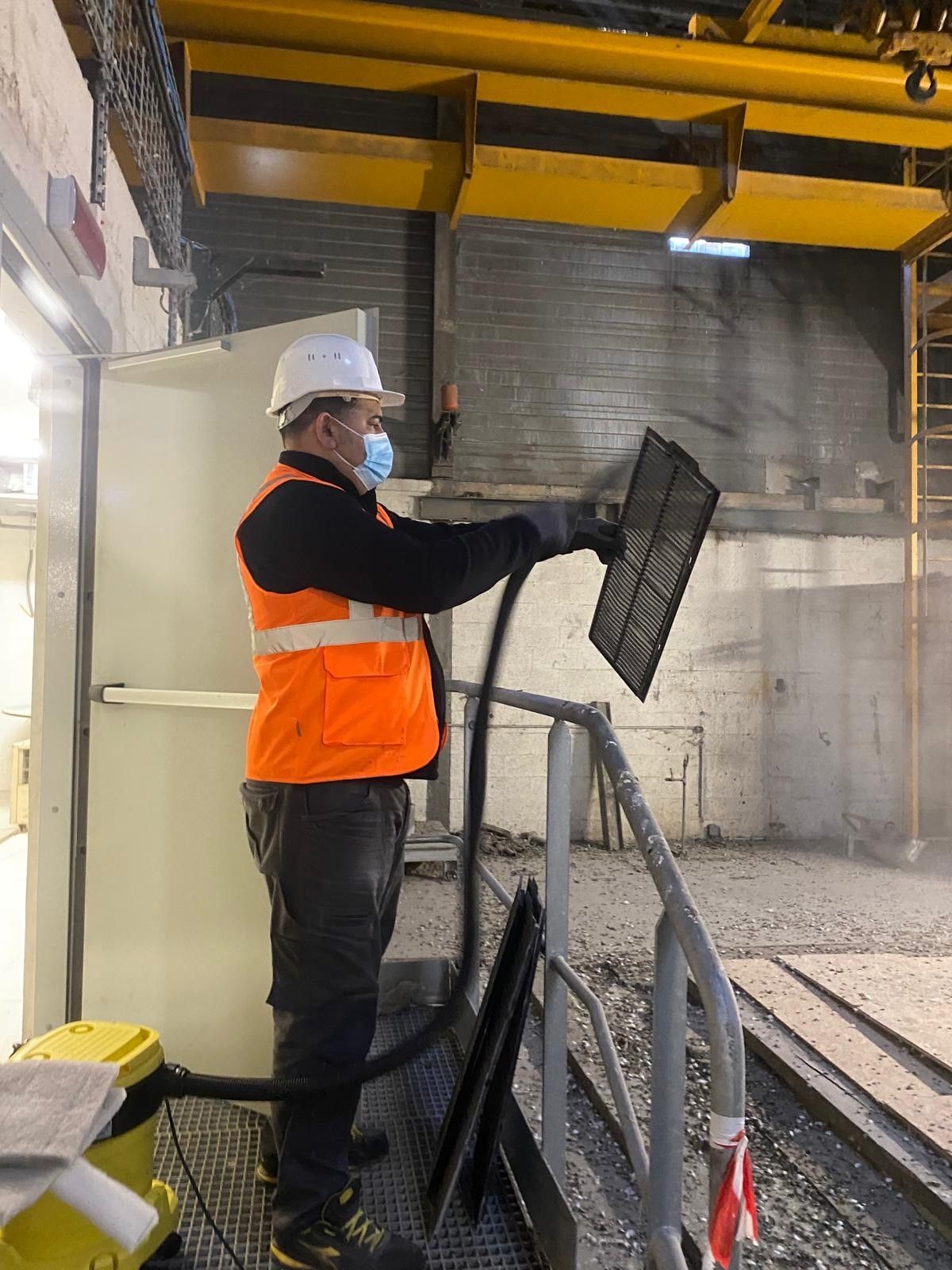 Un ouvrier portant un gilet orange utilise un tuyau d'aspirateur pour nettoyer un filtre dans un entrepôt.