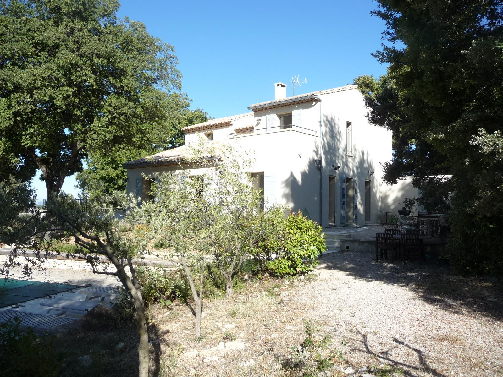 Maison blanche au toit de tuiles, dans un jardin ensoleillé, entourée d'arbres et d'arbustes.