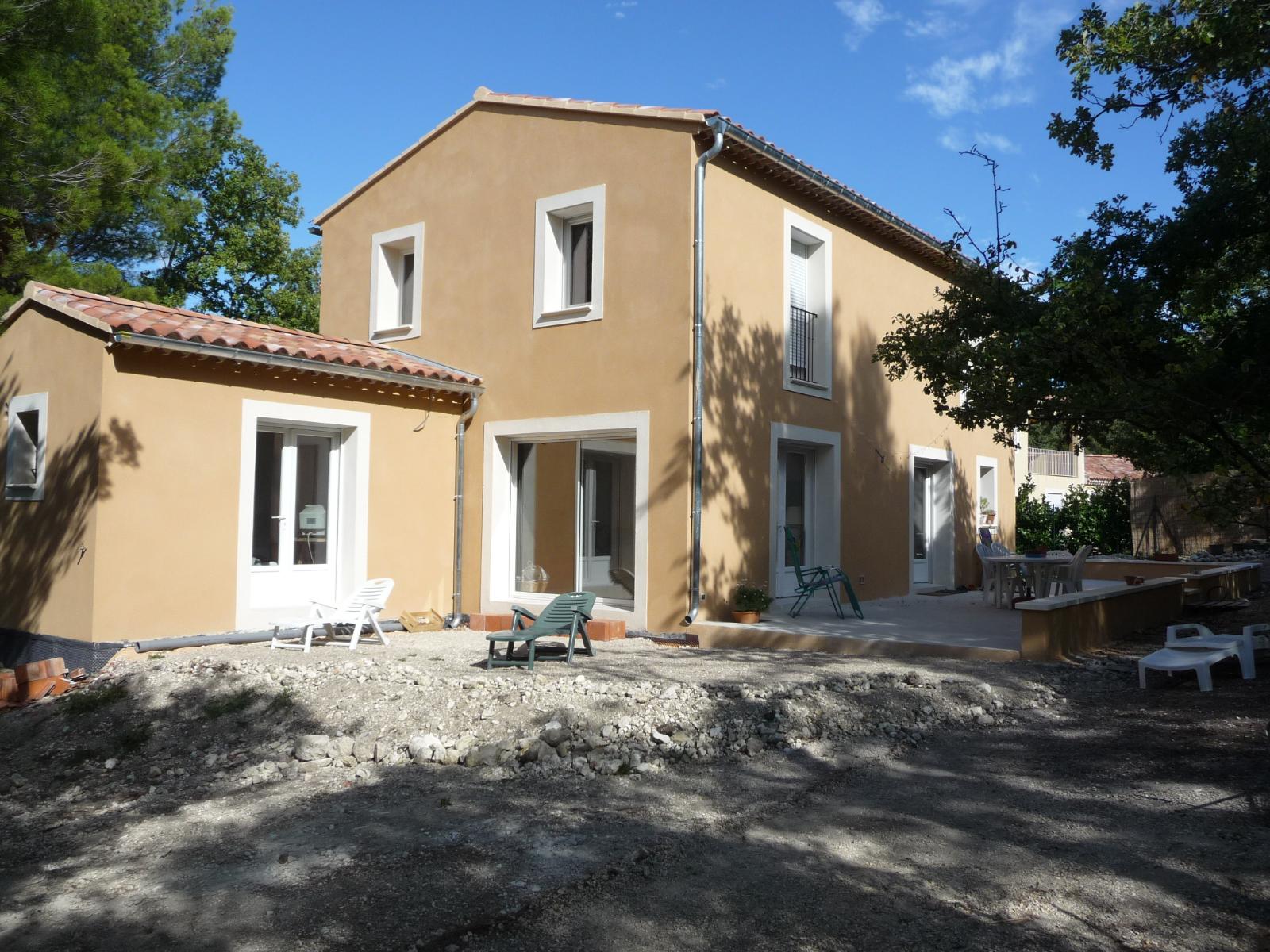 Maison en stuc beige avec des boiseries blanches et une cour en gravier sous un ciel bleu clair