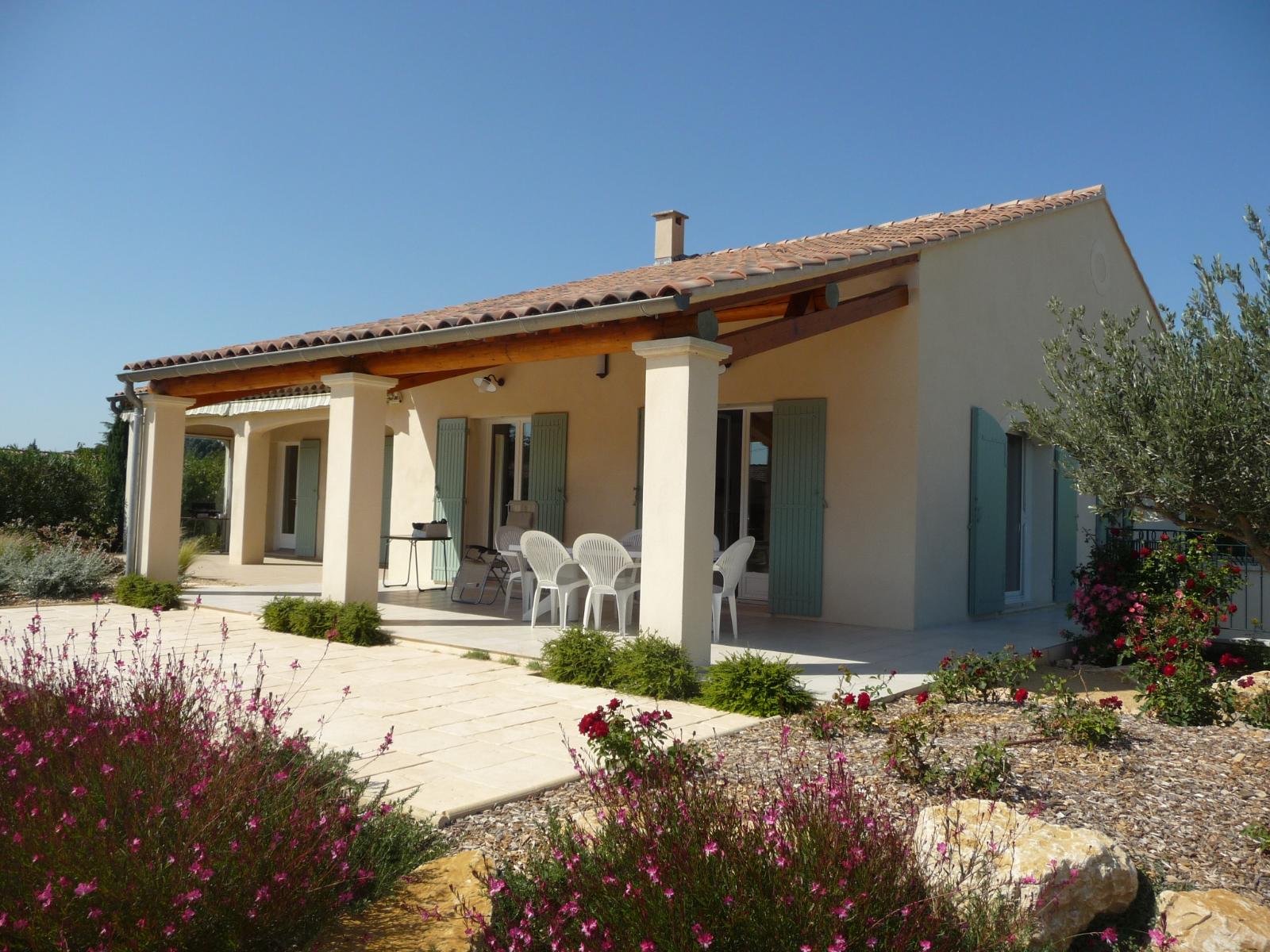 Maison moderne couleur crème avec un toit de tuiles, des chaises de patio et des fleurs de jardin sous un ciel bleu clair
