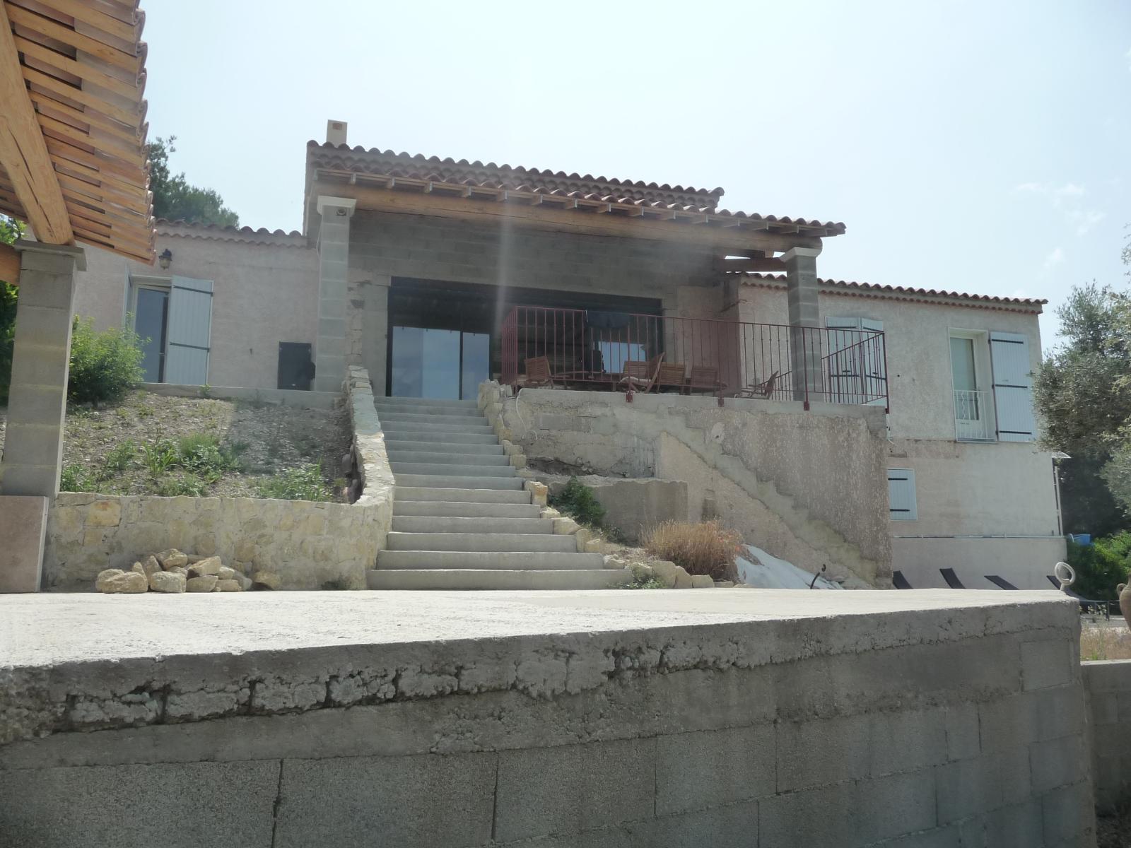 Maison en pierre avec un escalier central et un porche, située à flanc de colline et baignée de lumière naturelle.