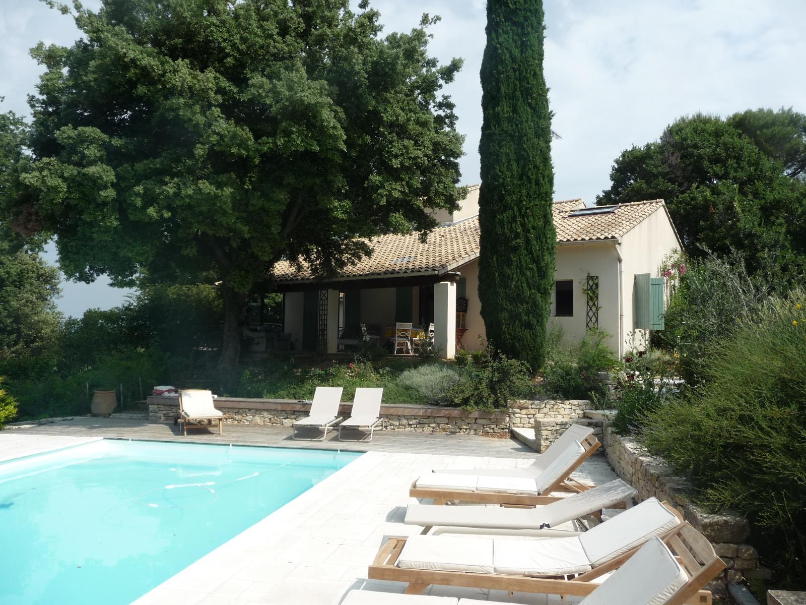 Villa au bord de la piscine avec chaises longues blanches, eau turquoise et végétation luxuriante sous de grands arbres
