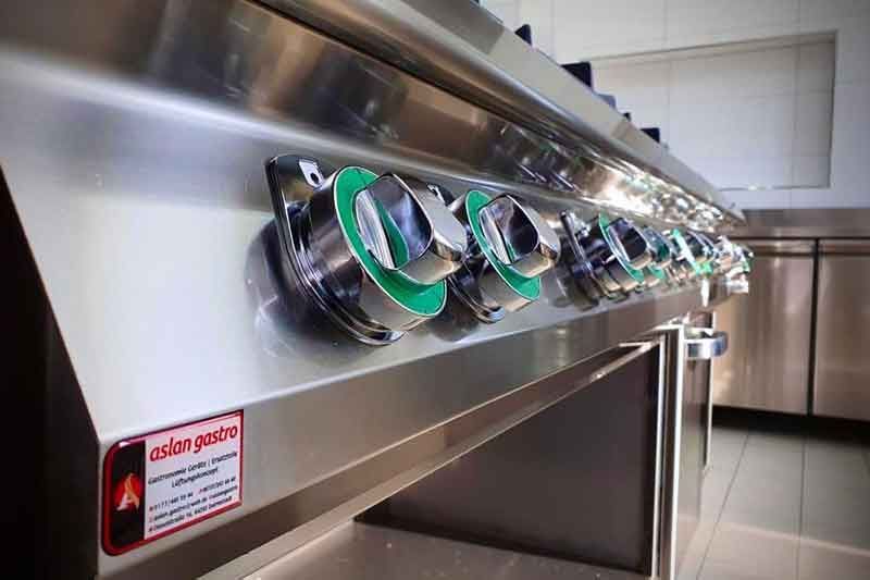 Kommerzieller Gasherd aus Edelstahl mit sechs Bedienknöpfen. Grüne Akzente auf den Knöpfen in einer Küche.