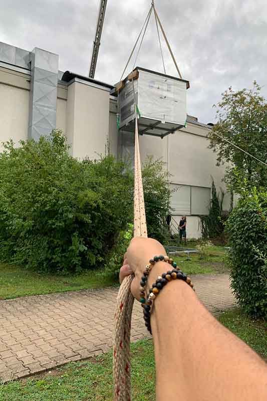 Der Arm einer Person hält ein Seil, eine HLK-Einheit wird von einem Kran in der Nähe eines Gebäudes angehoben.