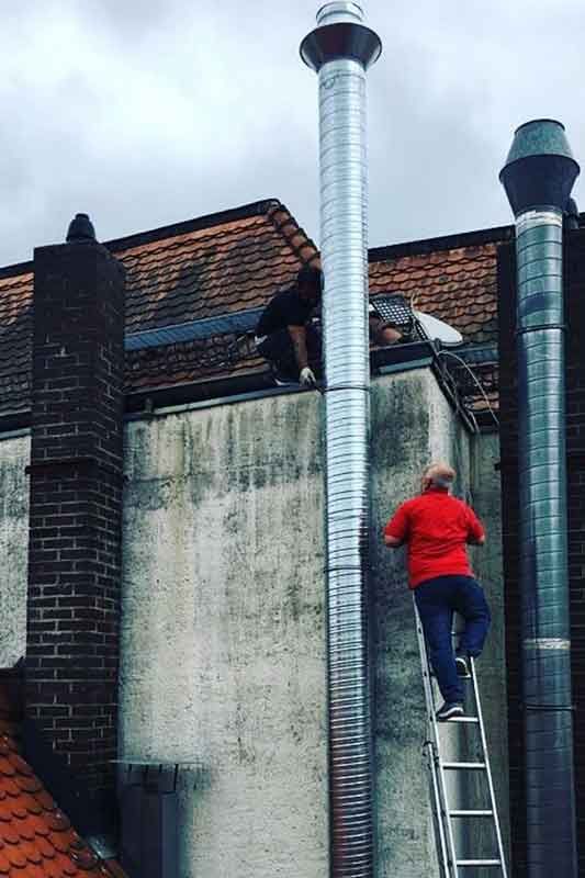 Zwei Personen auf einem Dach arbeiten an Schornsteinen. Einer klettert auf eine Leiter, der andere ist auf dem Dach.