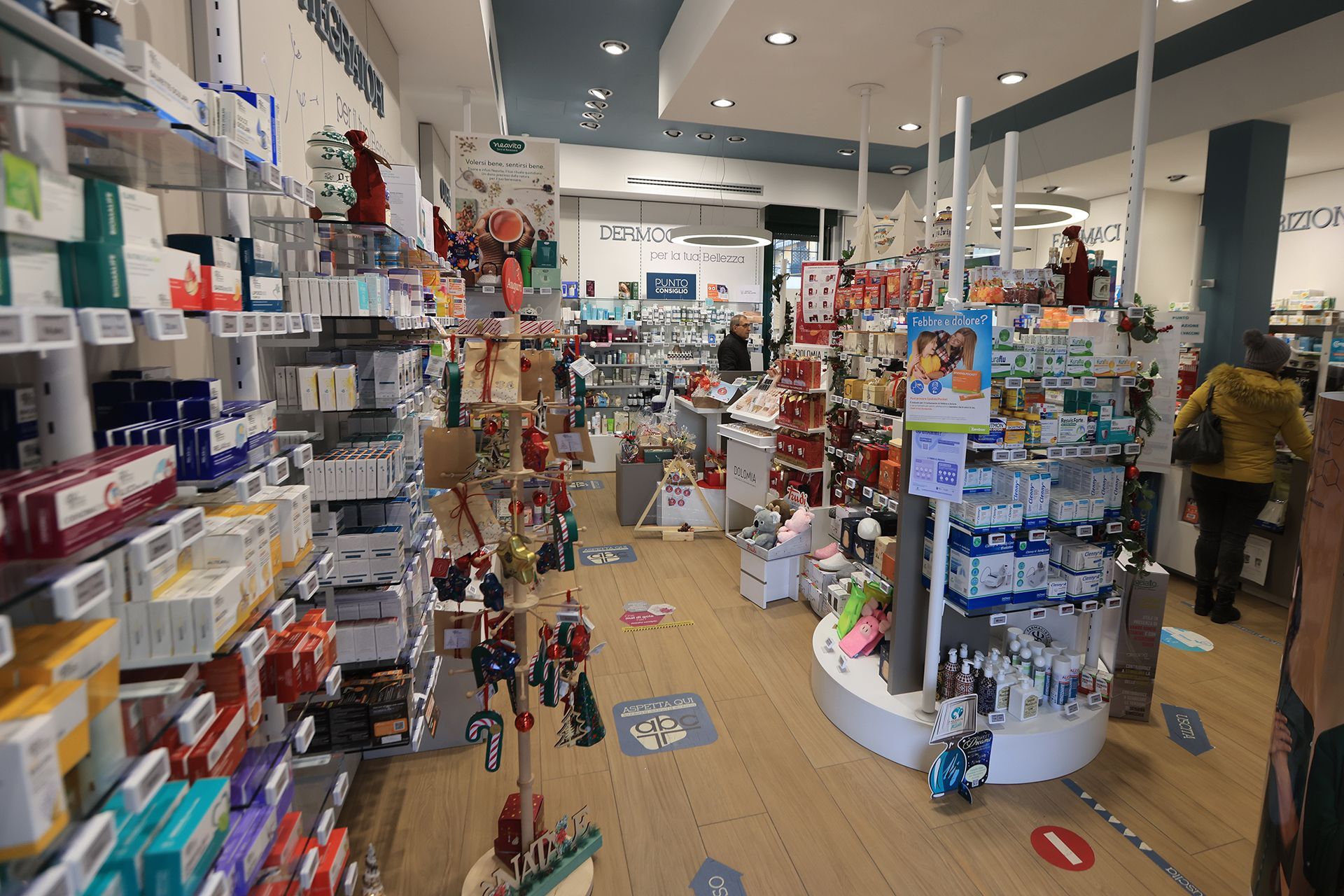 Interno della farmacia con scaffali pieni di prodotti e qualche cliente che curiosa.