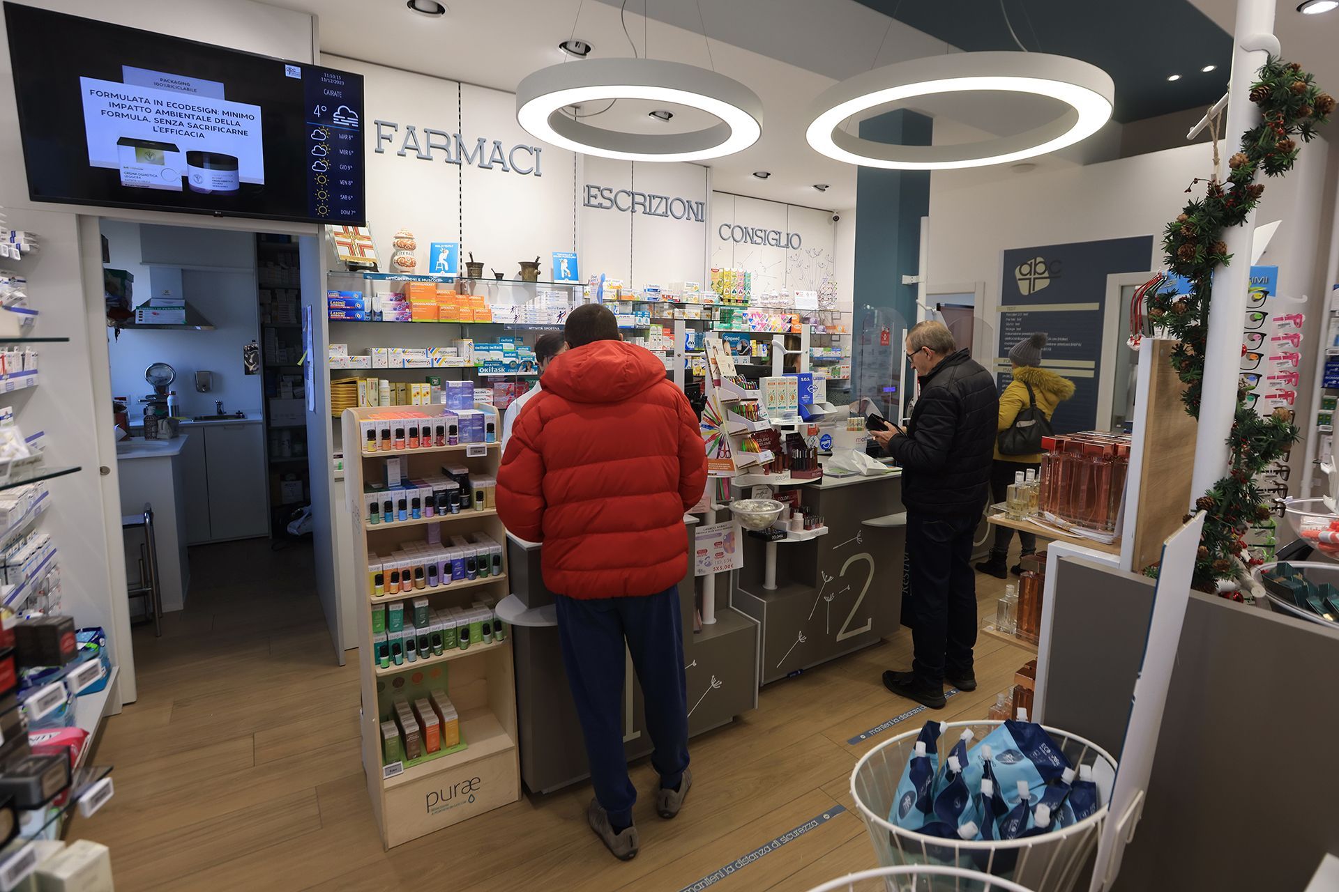 All'interno della farmacia, i clienti esaminano i prodotti e aspettano alla cassa. Gli scaffali sono pieni di merce.