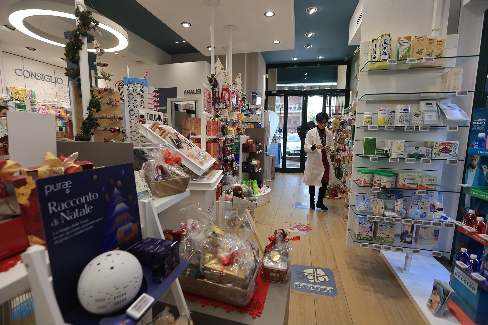 Interno della farmacia con una persona in camice, scaffali pieni di prodotti e cesti regalo.