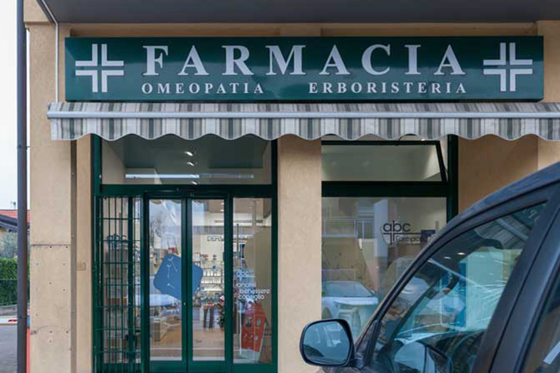 Vetrina della farmacia con insegna verde e tenda da sole, e un'auto parcheggiata di fronte.