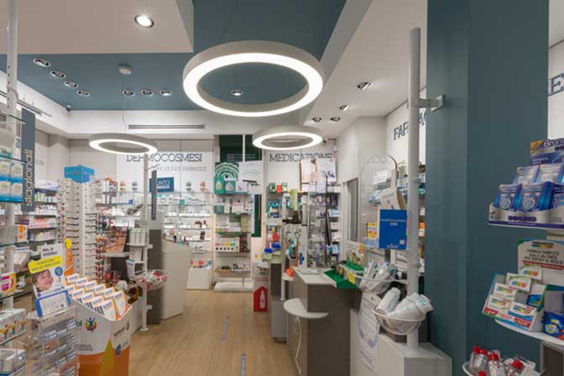 Interno della farmacia con pareti bianche e verde acqua, luci circolari e scaffali pieni di prodotti.