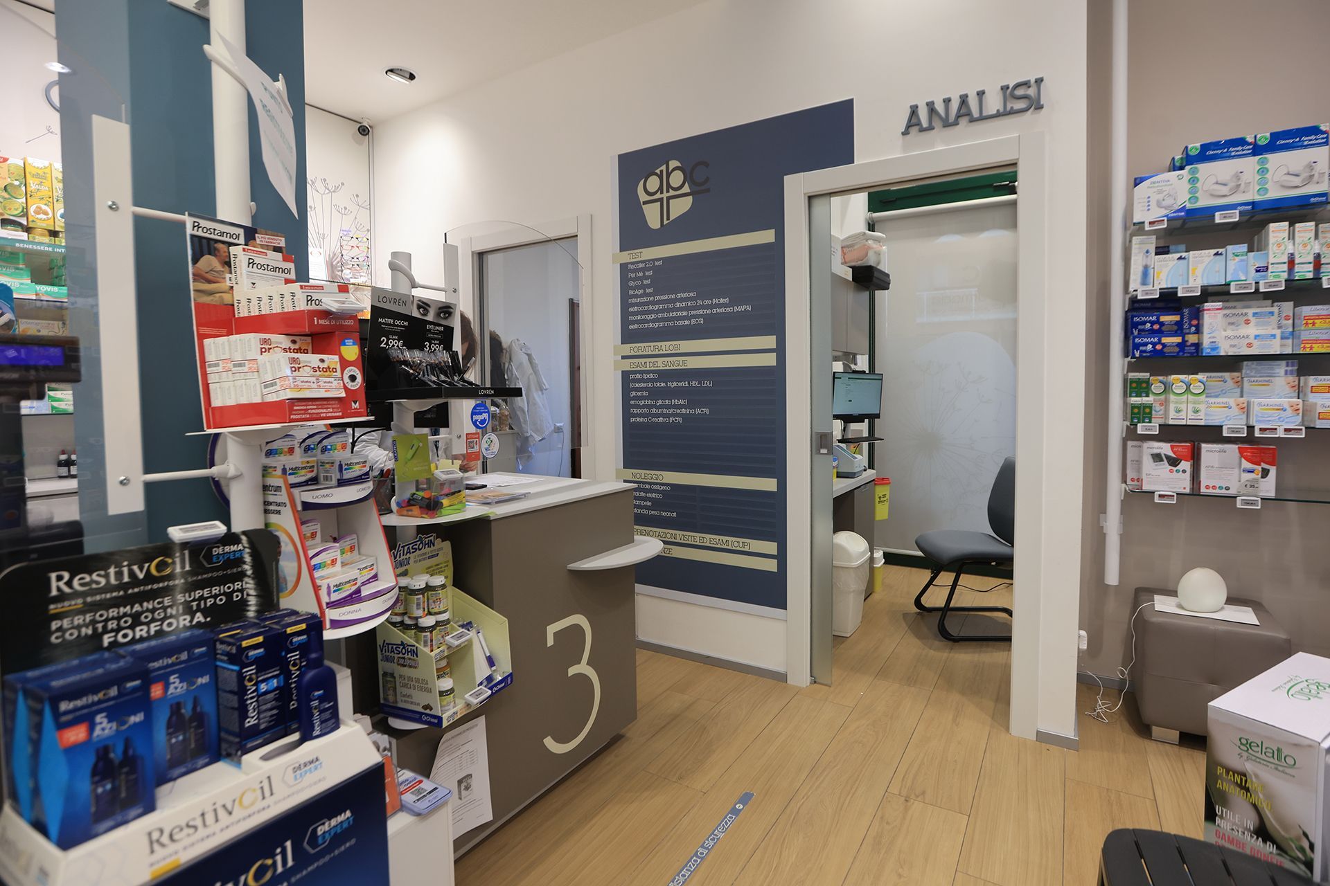 Interno della farmacia con scaffali pieni di prodotti, un bancone con il numero 3 e una porta con la scritta 