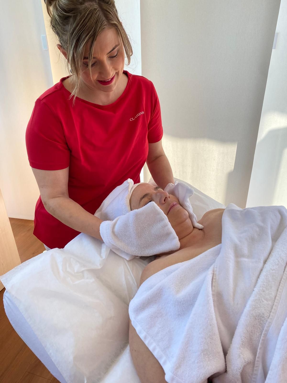 Eine Frau in rotem Hemd führt eine Gesichtsbehandlung an einer in ein weißes Handtuch gehüllten Kundin durch.