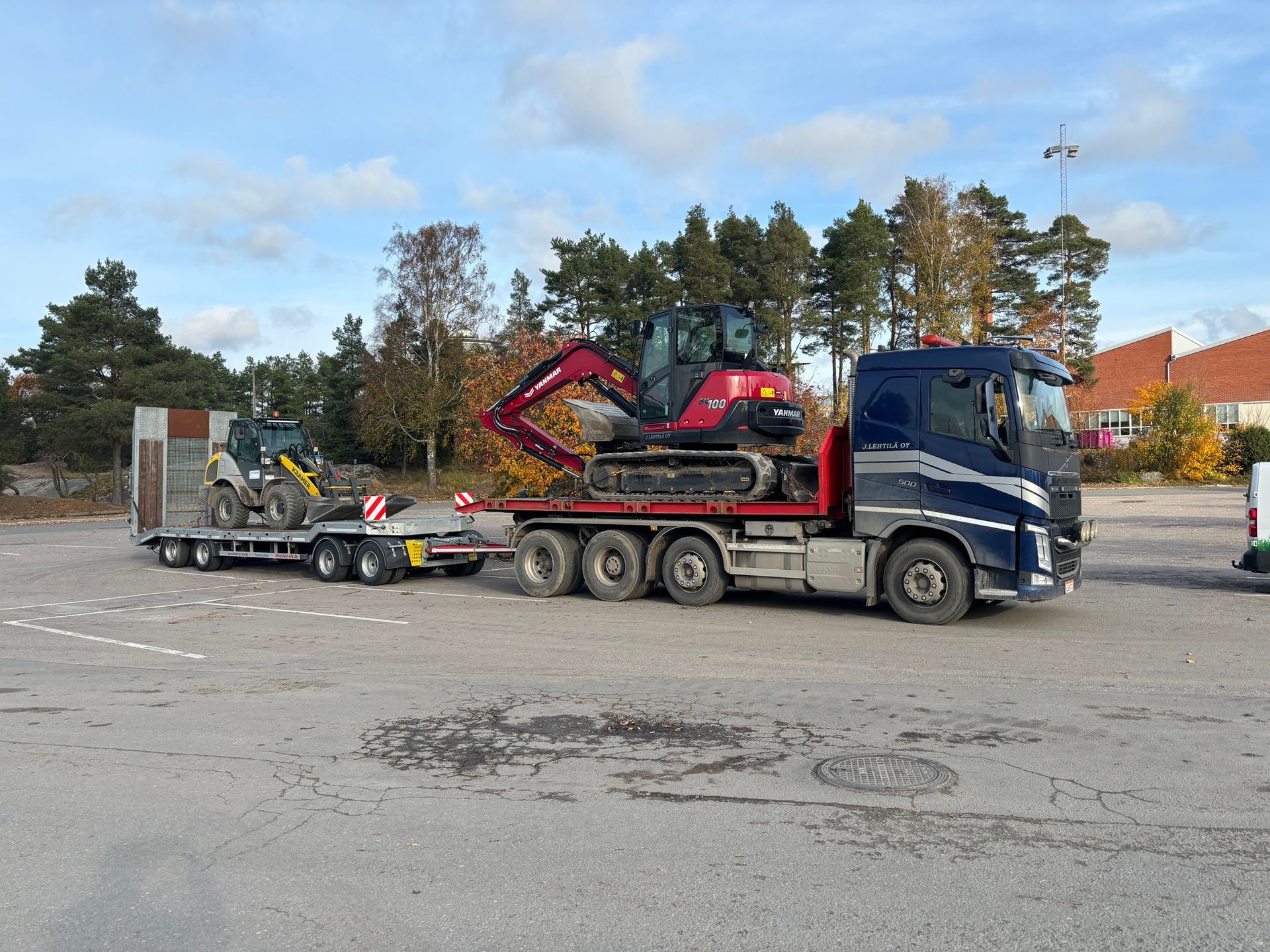 Lavettirekka kuljettaa punaista kaivinkonetta pysäköintialueella pilvisen taivaan alla