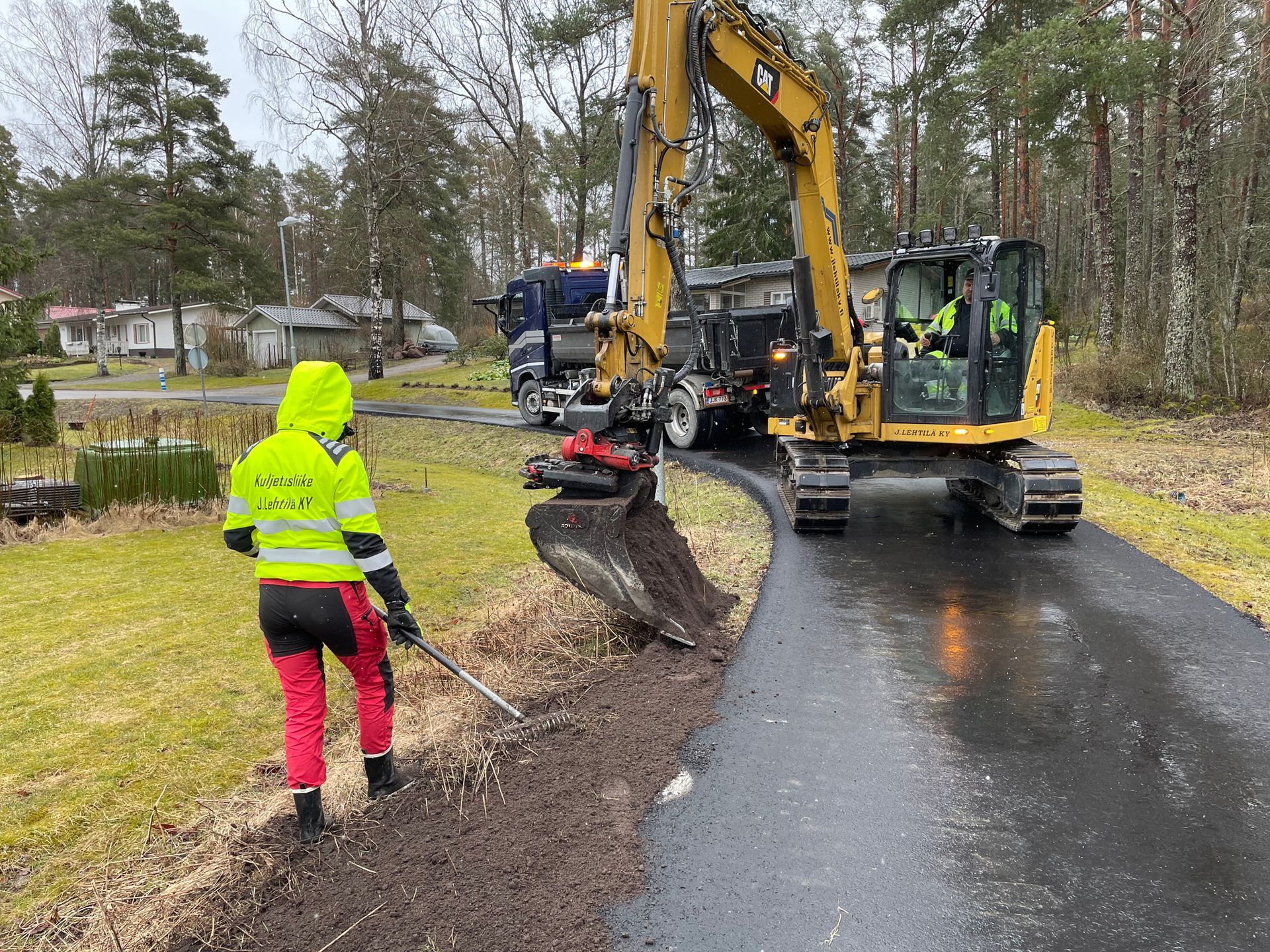 Neontakkiin pukeutunut työntekijä ohjaa kaivinkonetta, joka kaivaa märän tienvarren vieressä