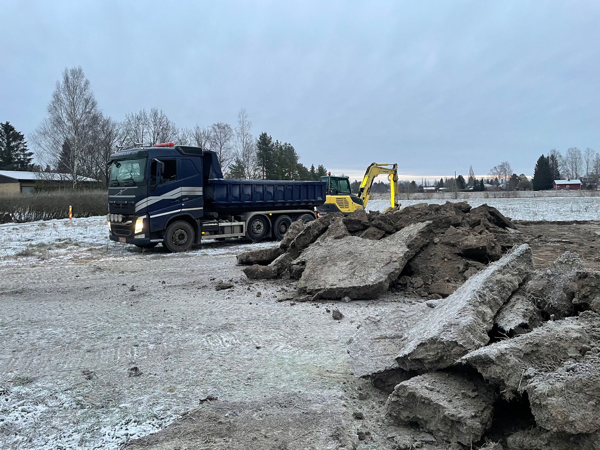 Sininen kippiauto ja keltainen kaivinkone betonimurskekasan vieressä lumisella työmaalla