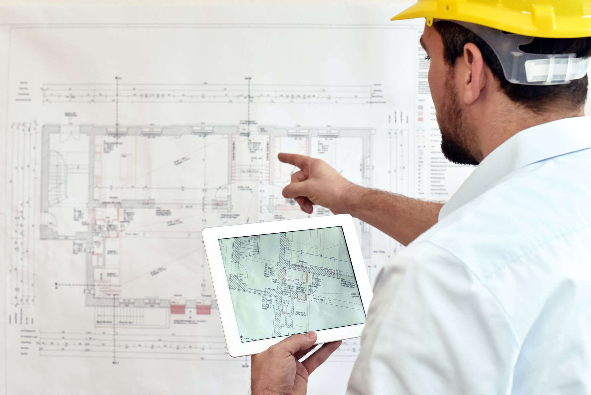 Un ouvrier du bâtiment pointe un plan et une tablette, portant un casque de sécurité.