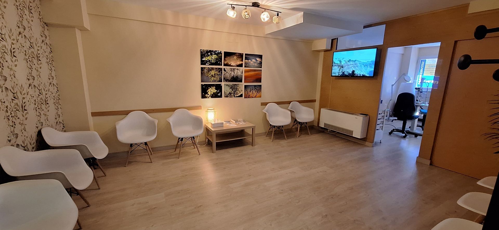 Una luminosa sala de espera con sillas blancas, una mesa pequeña, cuadros en las paredes y un televisor en una pared de color beige.