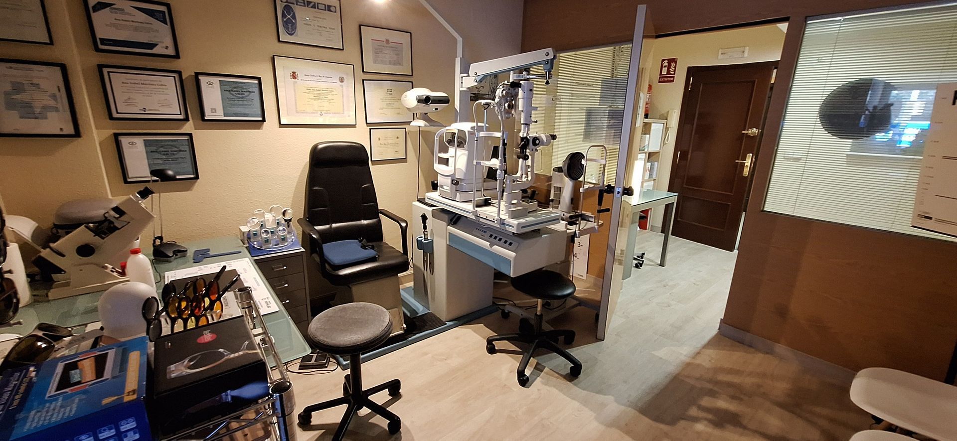 Un consultorio oftalmológico equipado con sillones de exploración médica y maquinaria de diagnóstico, con diplomas enmarcados en las paredes.