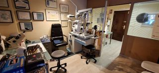 Un consultorio oftalmológico equipado con sillones de exploración médica y maquinaria de diagnóstico, con diplomas enmarcados en las paredes.