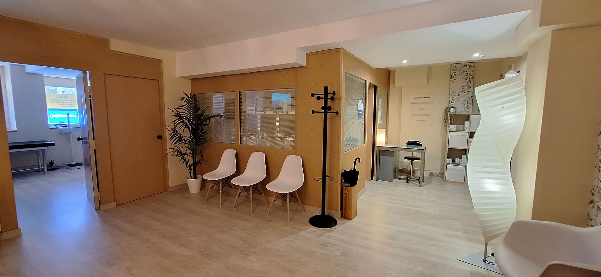 Una sala de espera con suelo de madera clara, tres sillas blancas, un perchero, una planta en maceta y una lámpara alta y ondulada.