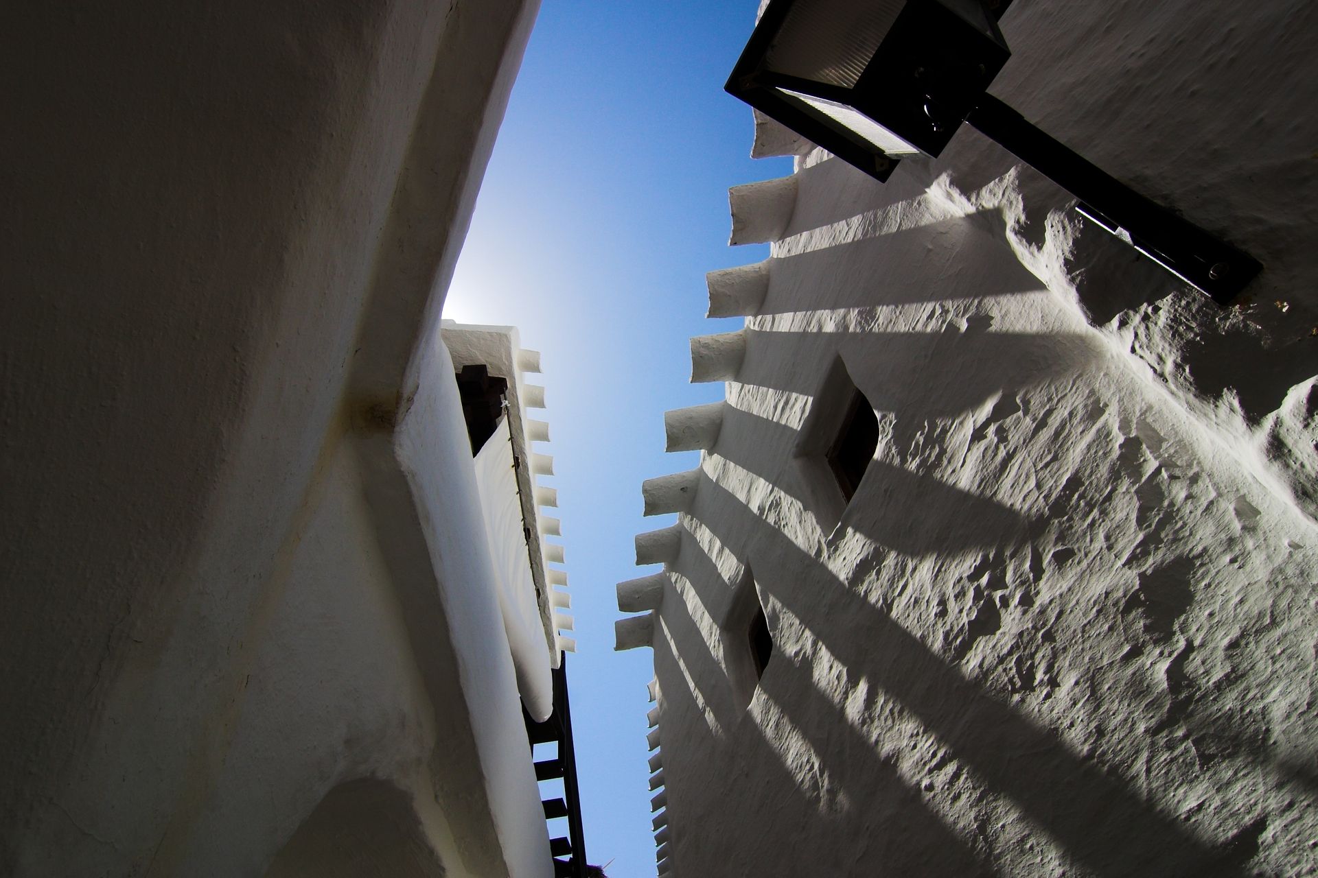 Mirando hacia arriba entre los edificios altos, se divisa un cielo azul brillante, con la intensa luz del sol proyectando sombras.