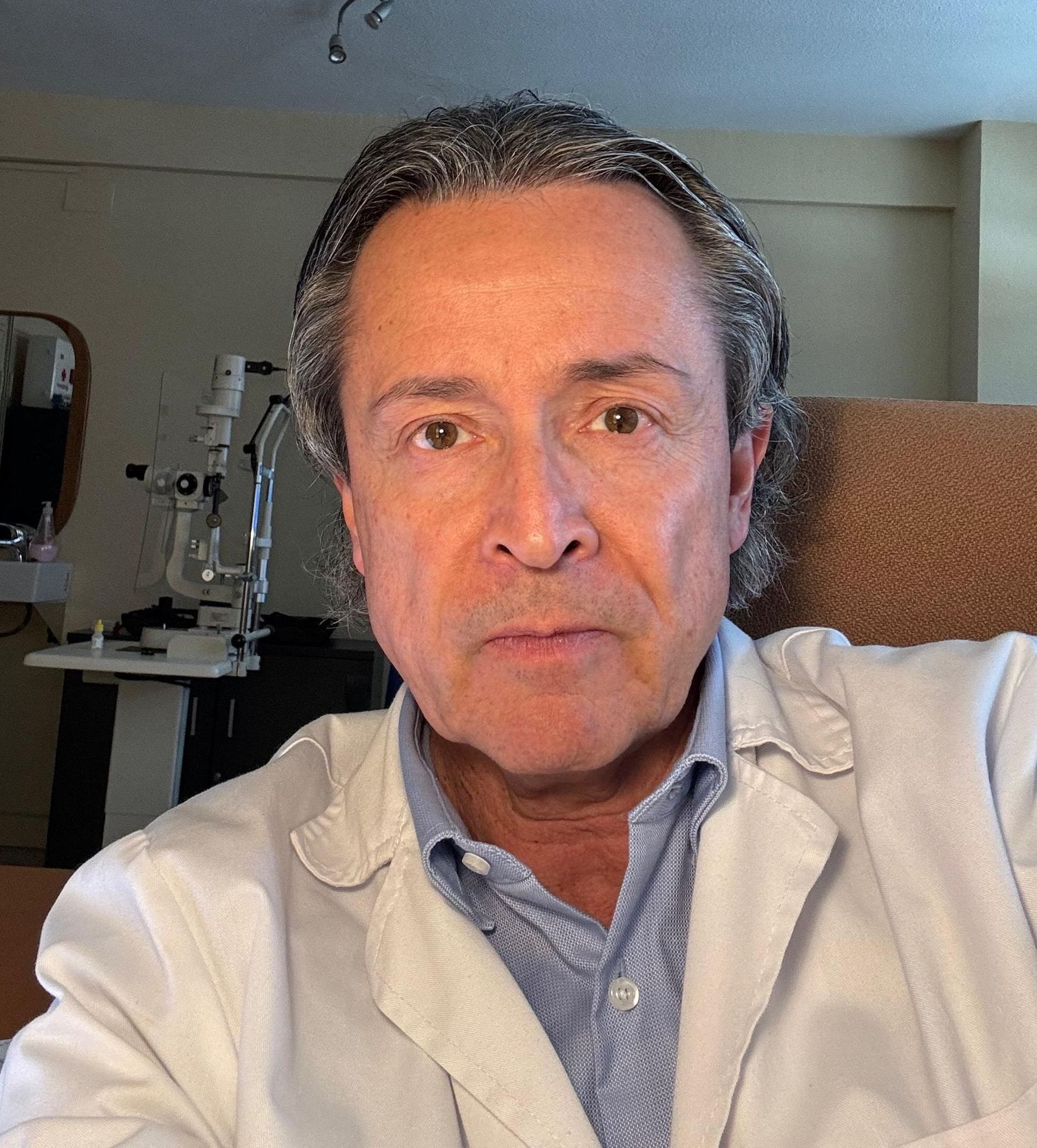 Una persona con bata blanca de laboratorio posando para una selfie en un consultorio clínico, con equipo médico visible al fondo.