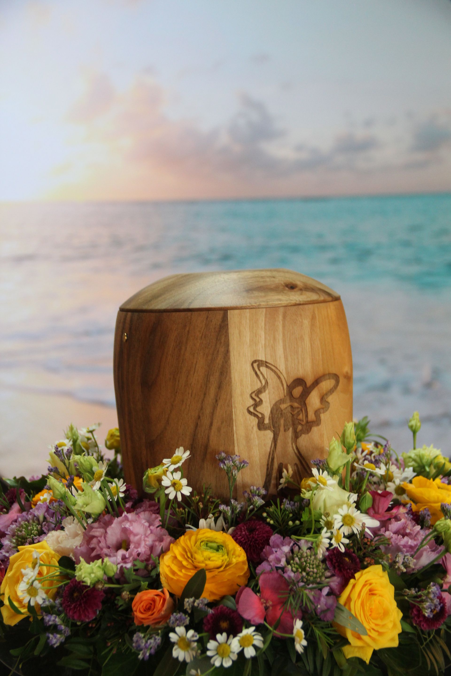 Eine hölzerne Urne, umgeben von bunten Blumen, steht am Strand bei Sonnenuntergang.