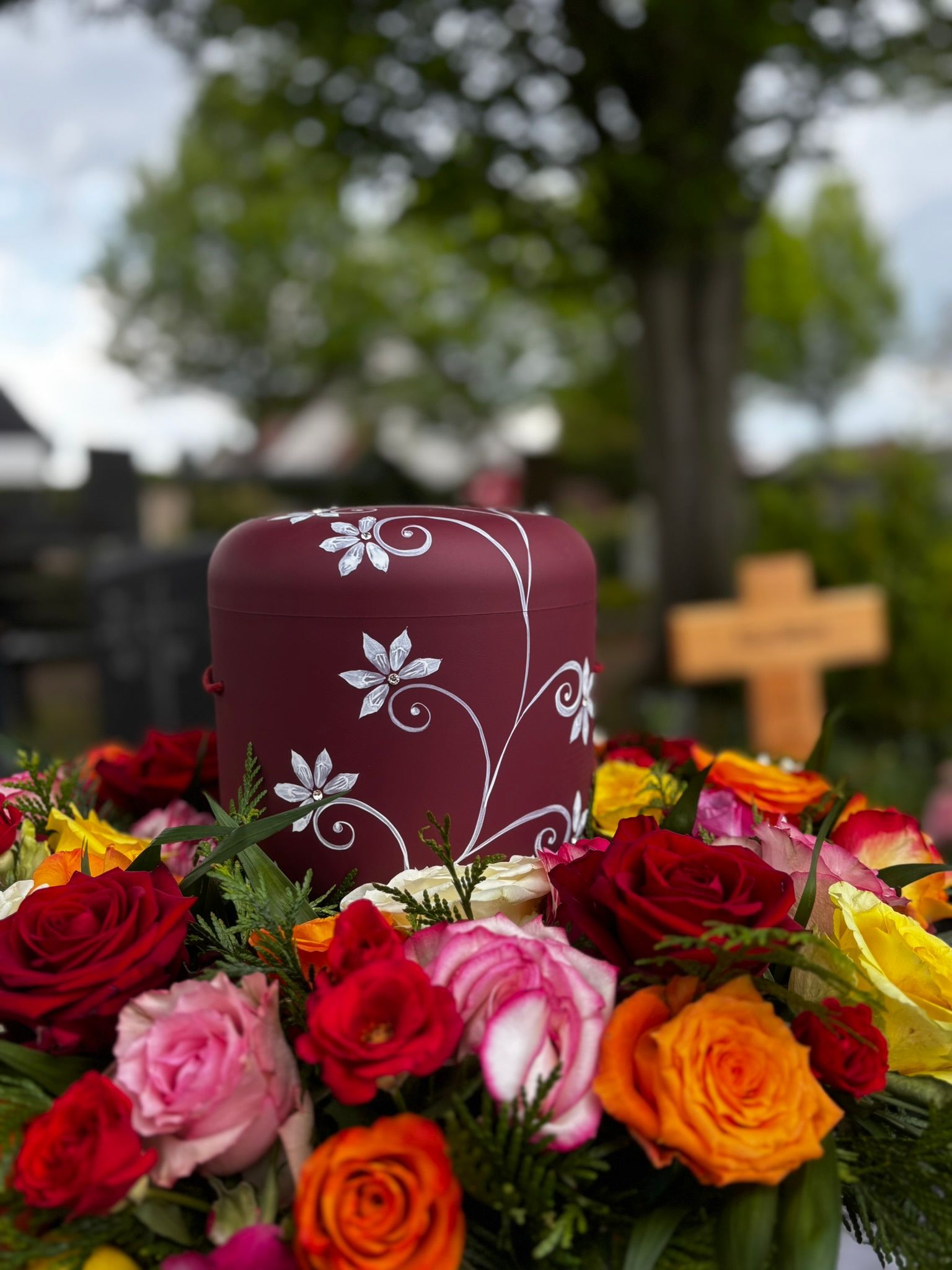 Eine bordeauxrote Blumenurne, umgeben von roten, rosa und orangefarbenen Rosen, in einer Friedhofsgedenkstätte