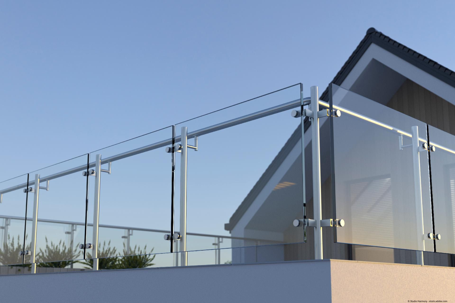 Moderner Glasbalkon mit Edelstahlbefestigungen an einer Hausfassade vor klarem blauen Himmel.