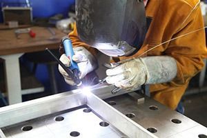 Person mit Schweißhelm und Schutzhandschuhen schweißt zwei Metallteile an einer Werkbank.