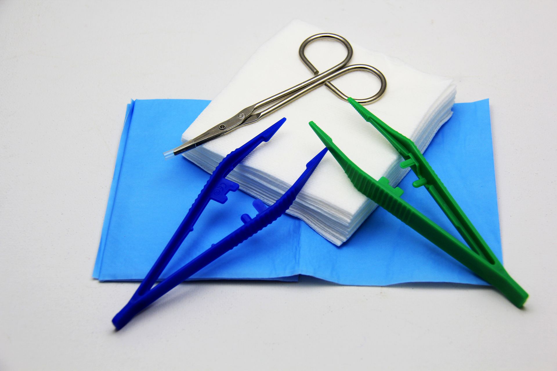 Pinces en plastique bleu et vert, ciseaux en métal, compresses de gaze et un champ opératoire bleu.