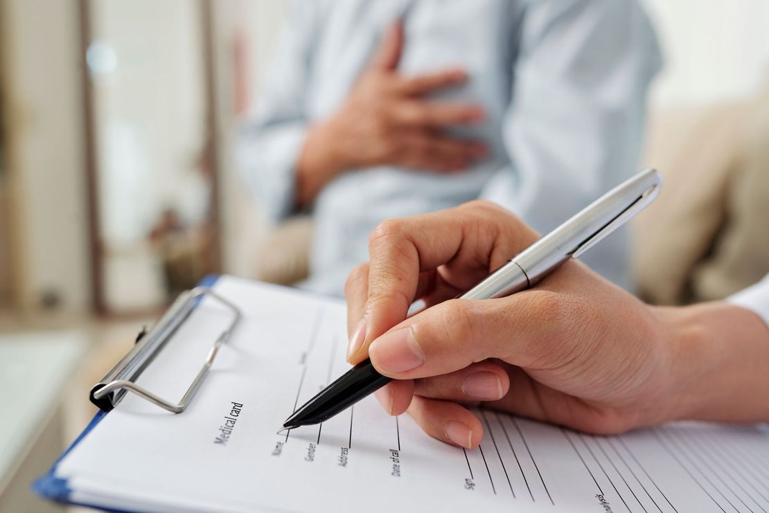 Un professionnel de santé prend des notes sur un formulaire tandis qu'un patient, en arrière-plan, se tient la poitrine.