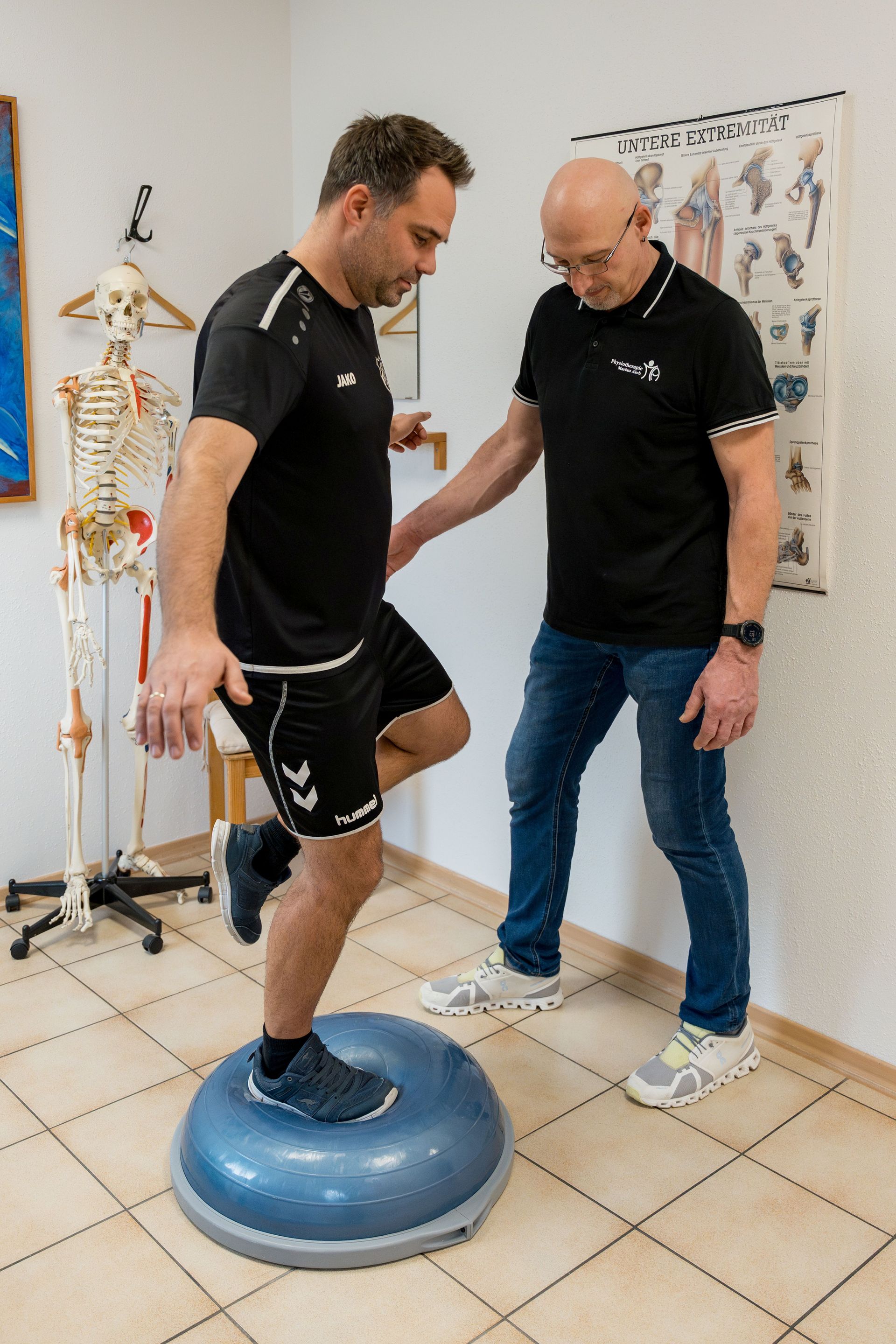 Ein Mann balanciert mit Unterstützung eines medizinischen Fachpersonals auf einem blauen Balancetrainer in einer Klinik.