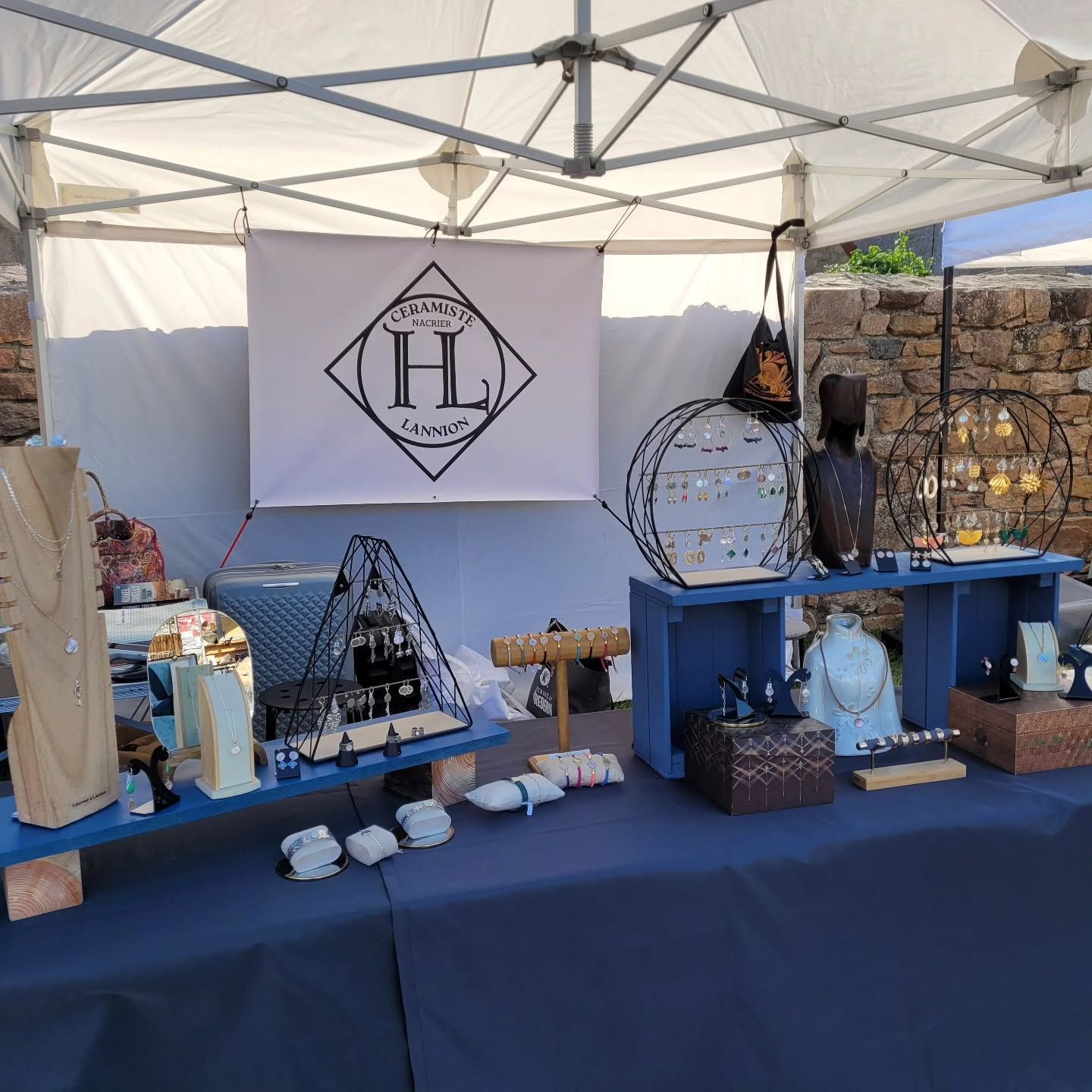 Stand d'artisanat extérieur avec nappe bleue, décorations faites main et banderole blanche sous un auvent.