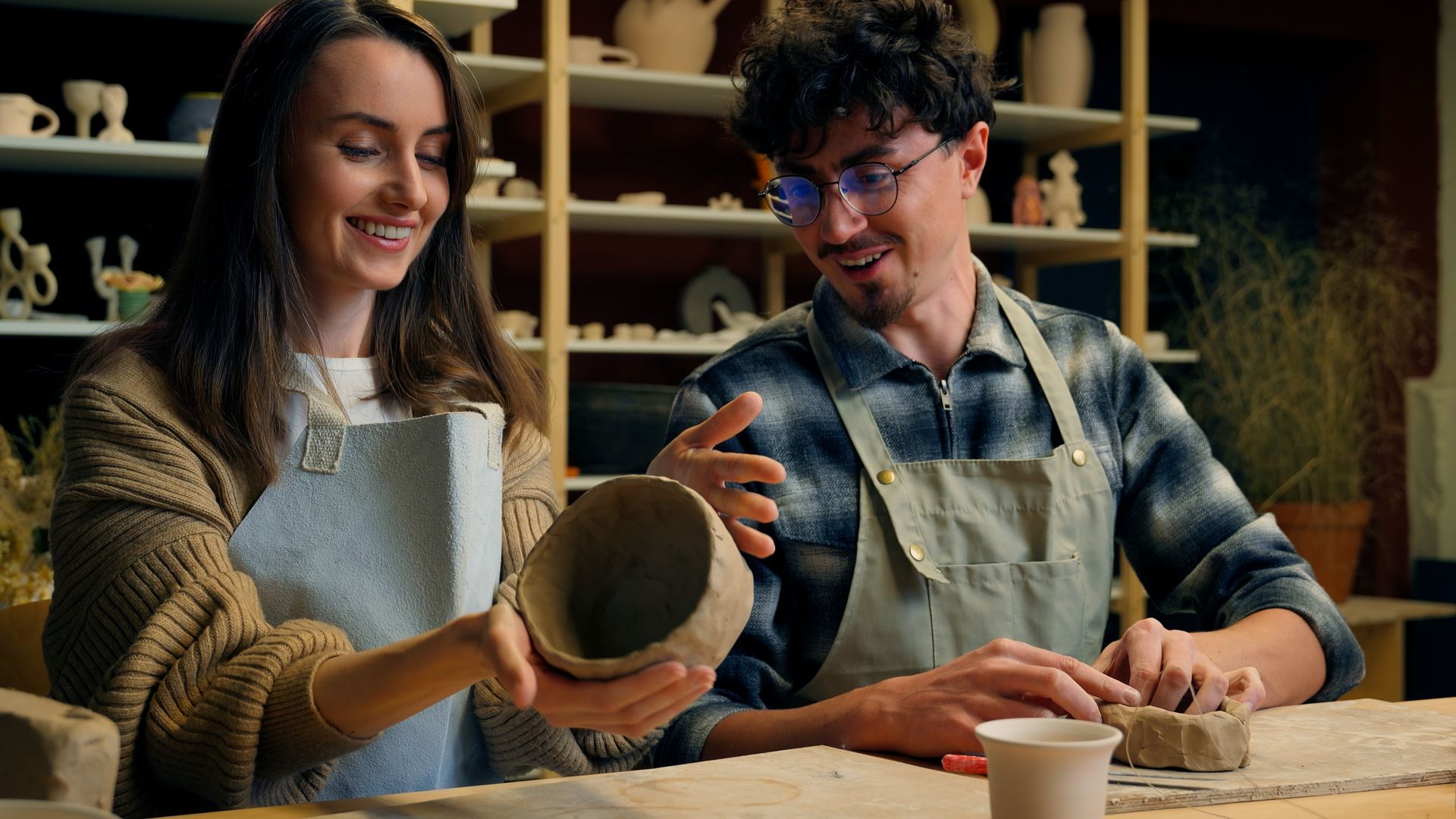 Deux personnes en tablier façonnent de la poterie à une table d'atelier, souriant devant un récipient en argile.