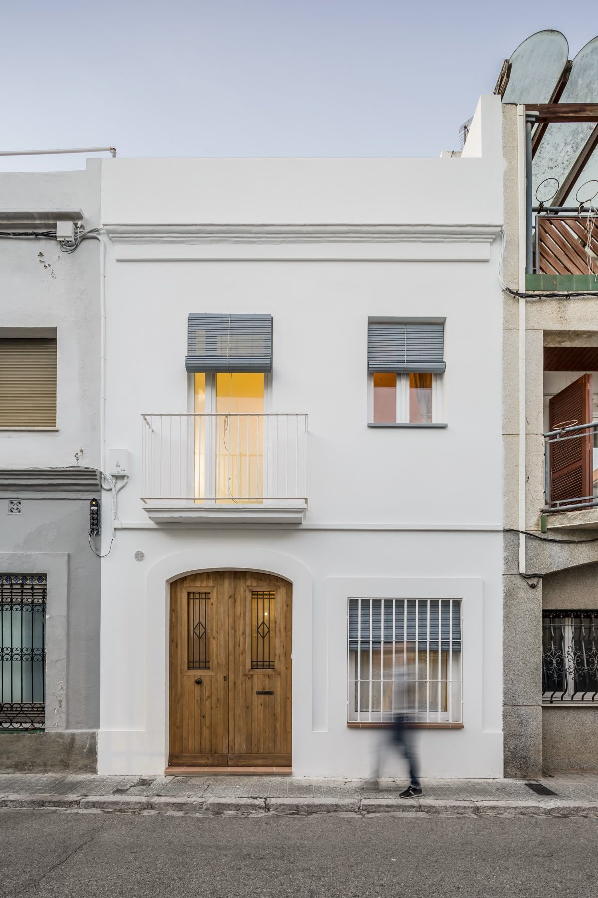 Edificio blanco de dos plantas con puerta de madera, balcón y ventanas con contraventanas. Una persona pasa.