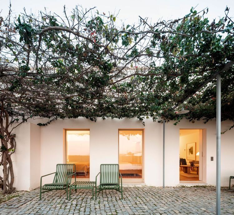Edificio blanco con tres puertas abiertas, sillas verdes y una pérgola cubierta de enredaderas. Patio adoquinado.