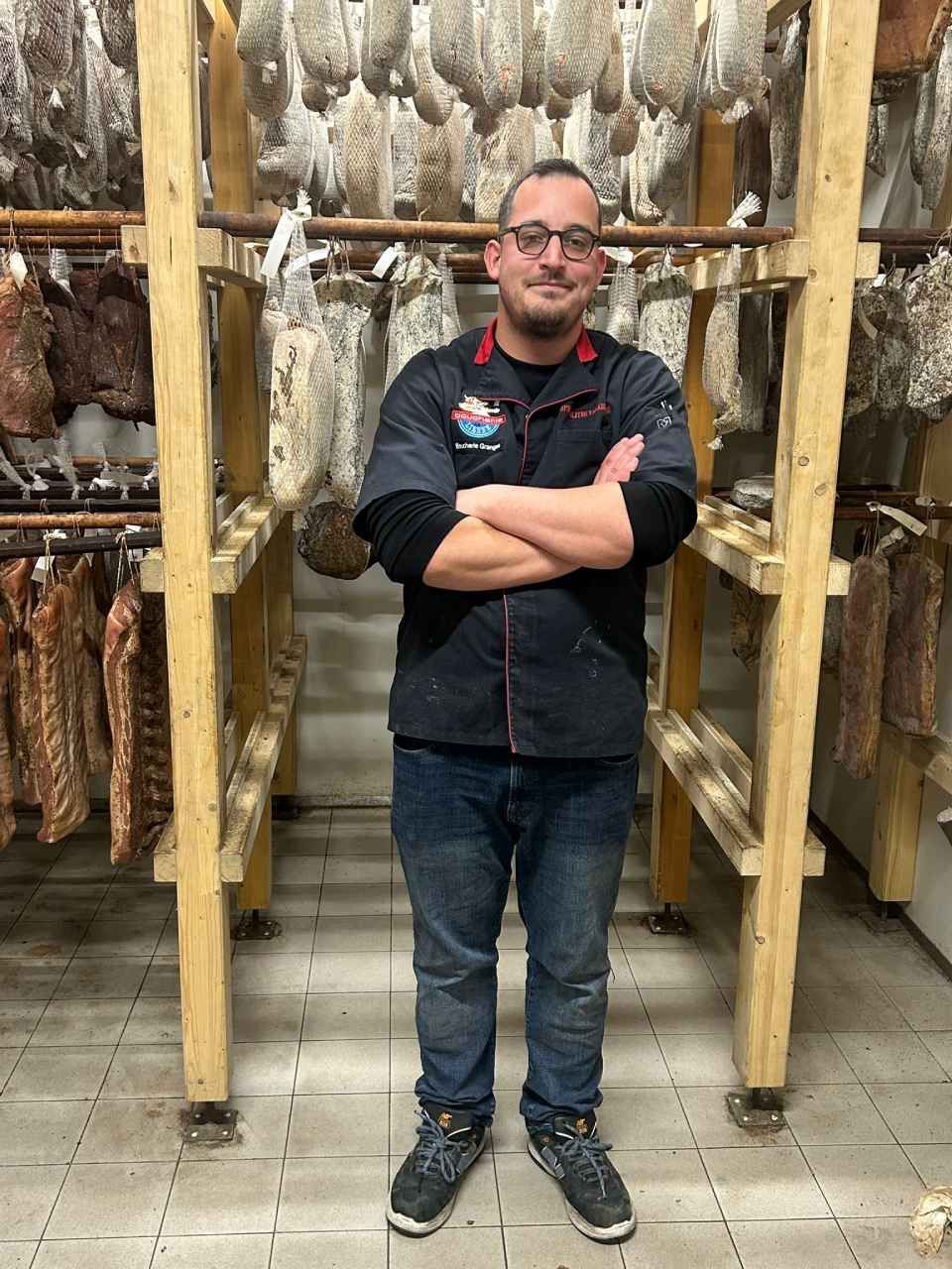 Un homme en chemise noire et jean se tient les bras croisés devant des charcuteries suspendues à des étagères en bois.