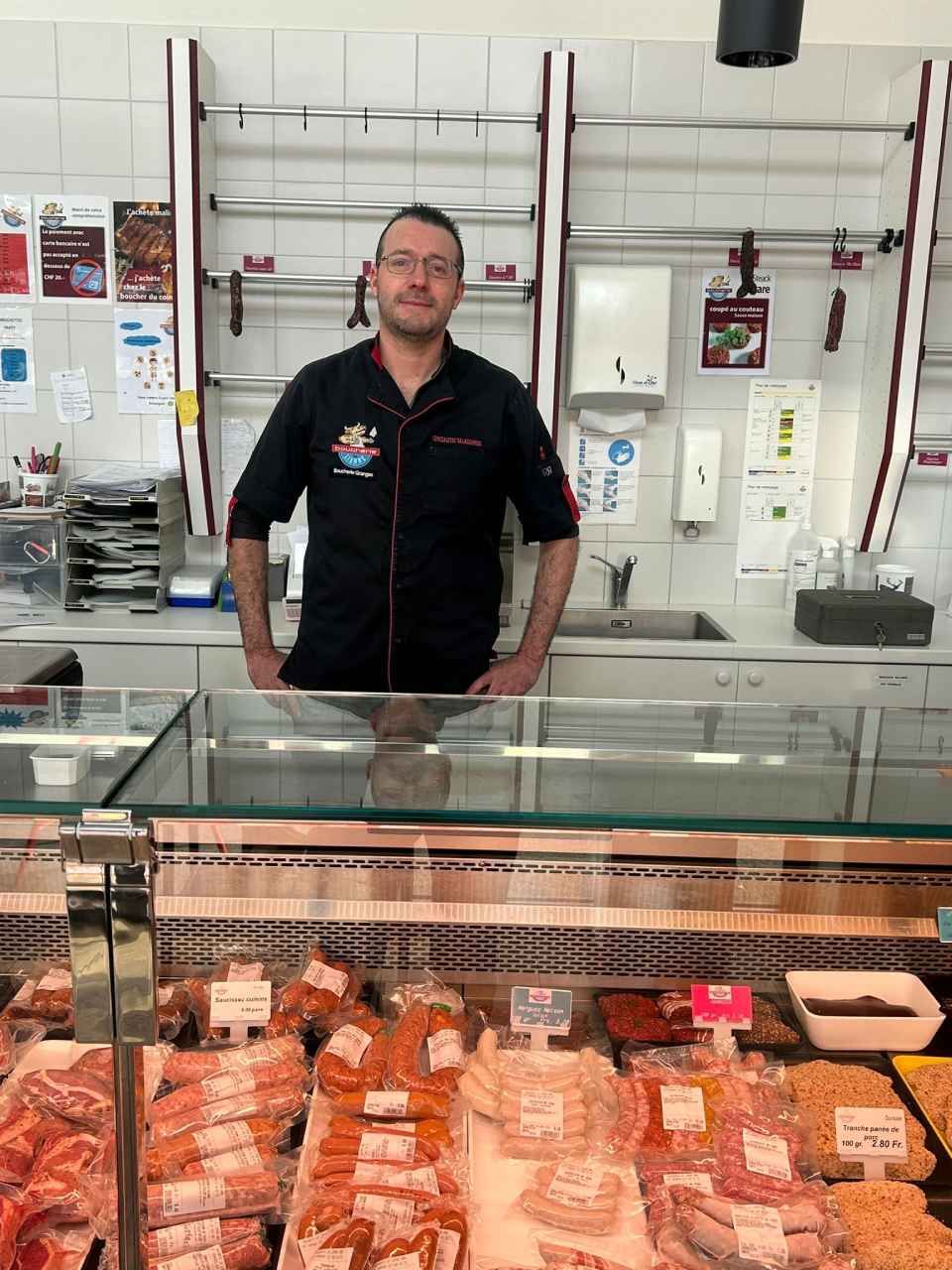 Un homme en uniforme de boucher noir se tient derrière une vitrine remplie de produits carnés.
